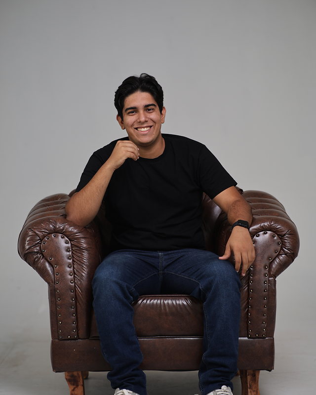 Victor Carvalho sentado em uma poltrona de couro marrom, de camiseta preta e calça jeans, sorrindo para a câmera.